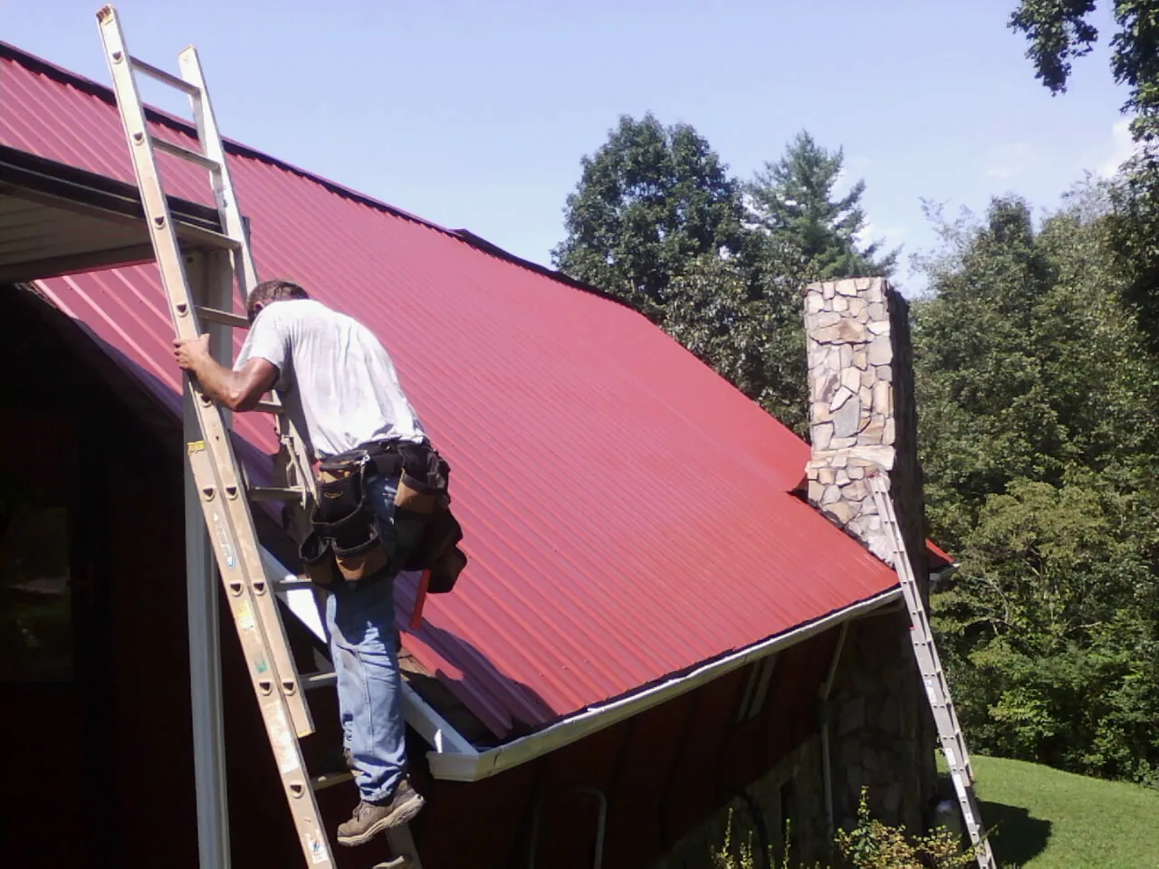 Roofer on a ladder beside a red metal roof and stone chimney — Soffit & Fascia Repair by Roofing Brothers in Timberville